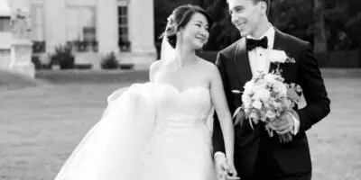 A black and white photograph of a bride and groom walking together on a lawn in front of a large stone building.
