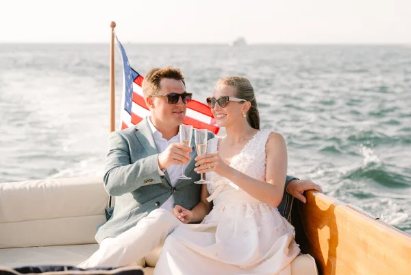 A bride and groom in formal wedding attire toasting with champagne glasses while seated on a boat on the water.
