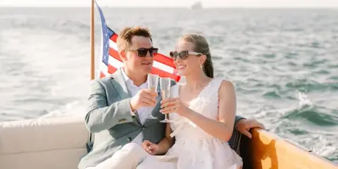 A bride and groom in formal wedding attire toasting with champagne glasses while seated on a boat on the water.