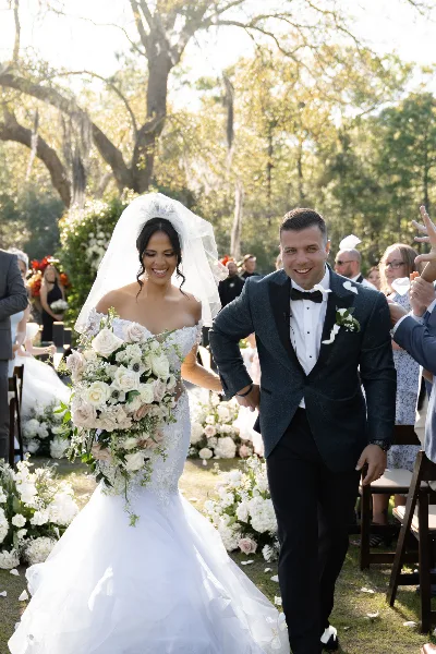 A bride and groom walking down a wedding aisle decorated with white flowers and greenery, with guests seated on either side.