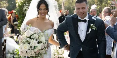 A bride and groom walking down a wedding aisle decorated with white flowers and greenery, with guests seated on either side.