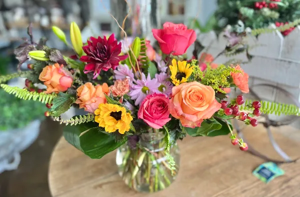 A colorful floral arrangement with orange roses, pink tulips, yellow sunflowers, and greenery in a glass vase.