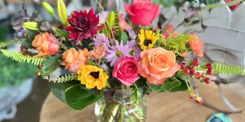 A colorful floral arrangement with orange roses, pink tulips, yellow sunflowers, and greenery in a glass vase.