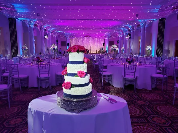 A wedding reception hall featuring a four-tier white and navy blue cake with pink roses, surrounded by set tables and purple ambient lighting.