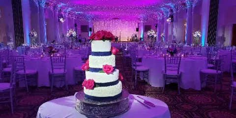 A wedding reception hall featuring a four-tier white and navy blue cake with pink roses, surrounded by set tables and purple ambient lighting.
