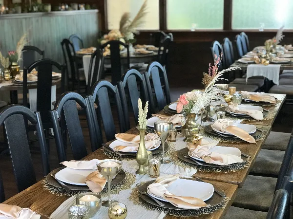 A long wooden banquet table set with white plates, folded napkins, metallic glassware, candles, and floral centerpieces in a restaurant dining room.