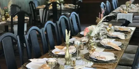 A long wooden banquet table set with white plates, folded napkins, metallic glassware, candles, and floral centerpieces in a restaurant dining room.
