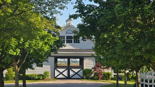 Exterior view of a white barn wedding venue with a cupola, black double doors, and white picket fencing surrounded by green trees.