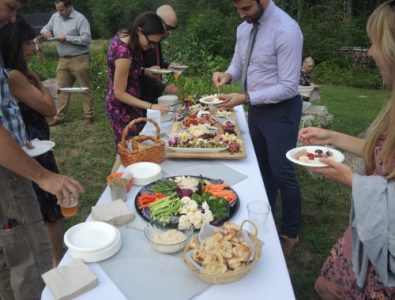 Guests serving themselves from an outdoor buffet table featuring vegetables, dips, and appetizers.
