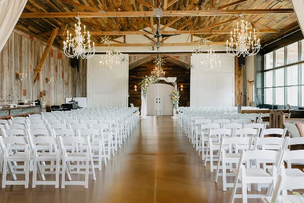 Interior of a rustic barn wedding venue with white chairs arranged for a ceremony, featuring wooden beams and chandeliers.