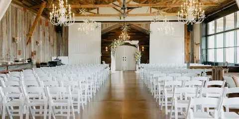 Interior of a rustic barn wedding venue with white chairs arranged for a ceremony, featuring wooden beams and chandeliers.