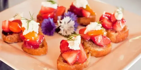A plate of gourmet canapés topped with fresh fruit, cream, and herbs, garnished with edible flowers.