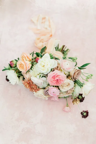 A close-up photograph of a floral arrangement with pink, white, and peach roses and ranunculus on a textured pink background.