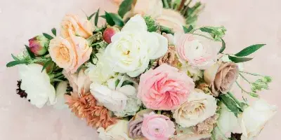 A close-up photograph of a floral arrangement with pink, white, and peach roses and ranunculus on a textured pink background.