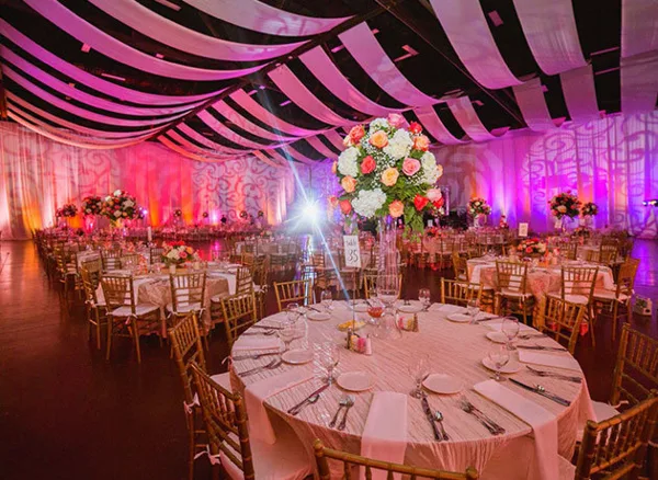 An indoor wedding reception venue featuring round tables, Chiavari chairs, and a large floral centerpiece under a draped ceiling.