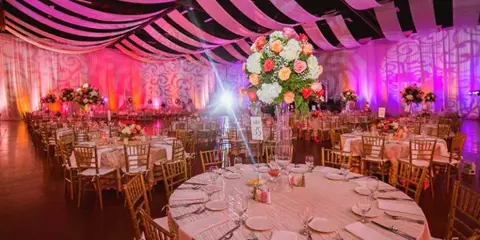 An indoor wedding reception venue featuring round tables, Chiavari chairs, and a large floral centerpiece under a draped ceiling.