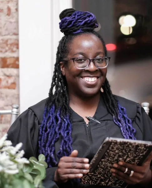 A smiling woman with purple-tipped dreadlocks and glasses holding a book, standing in front of a brick wall.