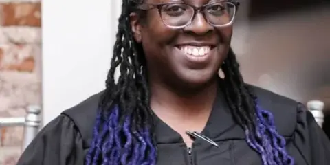A smiling woman with purple-tipped dreadlocks and glasses holding a book, standing in front of a brick wall.