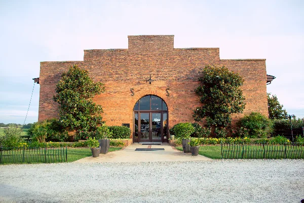 Exterior view of a large red brick barn with an arched glass entrance, flanked by green trees and a paved walkway.