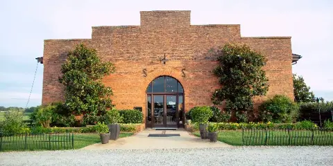 Exterior view of a large red brick barn with an arched glass entrance, flanked by green trees and a paved walkway.