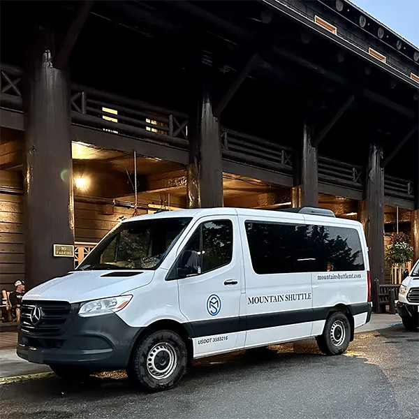 A white Mercedes-Benz Sprinter van with Mountain Shuttle branding parked in front of a wooden building.