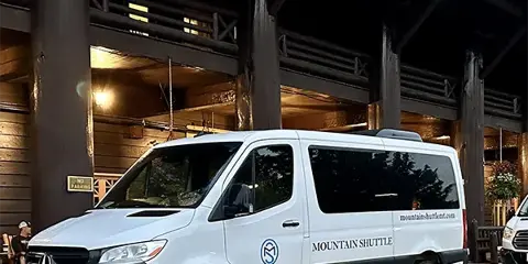 A white Mercedes-Benz Sprinter van with Mountain Shuttle branding parked in front of a wooden building.