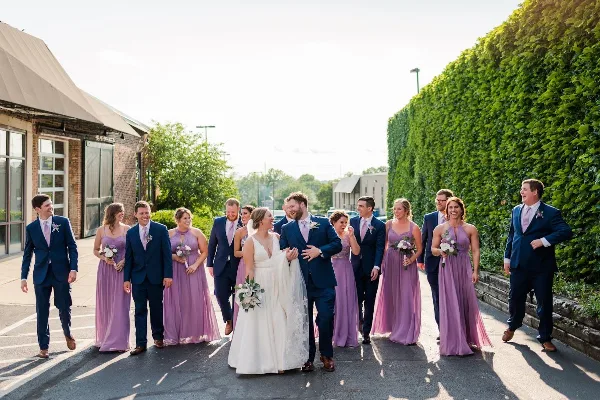 A wedding party including the bride, groom, and bridesmaids in purple dresses walking outdoors near a brick building and a tall green hedge.