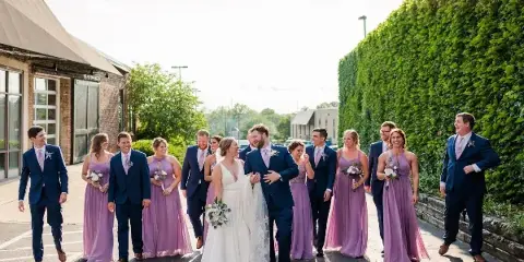 A wedding party including the bride, groom, and bridesmaids in purple dresses walking outdoors near a brick building and a tall green hedge.