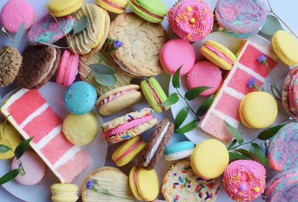 A colorful overhead arrangement of assorted baked goods including macarons, cookies, cupcakes, and slices of pink layer cake.