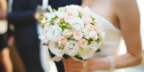 A close-up of a bride holding a round bouquet of white and blush roses with green accents.