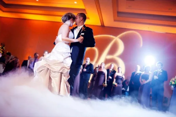 A bride and groom sharing a first dance on a stage with low-lying fog and warm orange lighting during a wedding reception.