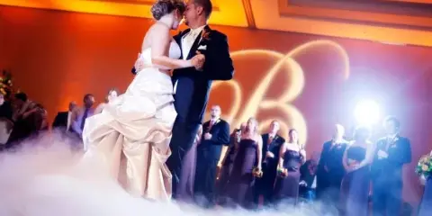 A bride and groom sharing a first dance on a stage with low-lying fog and warm orange lighting during a wedding reception.