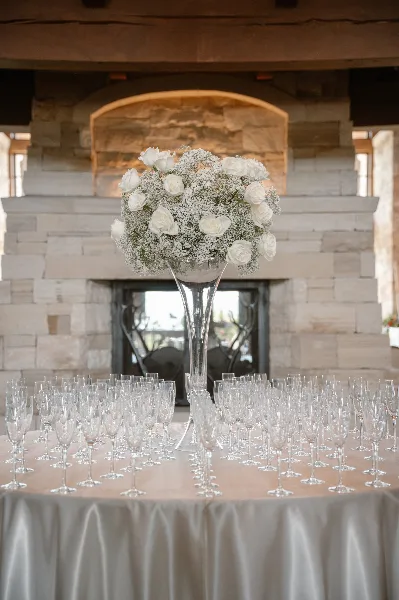A wedding table setting featuring a tall glass vase with white roses and baby's breath, surrounded by champagne flutes on a beige tablecloth.