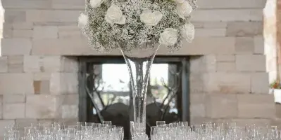 A wedding table setting featuring a tall glass vase with white roses and baby's breath, surrounded by champagne flutes on a beige tablecloth.