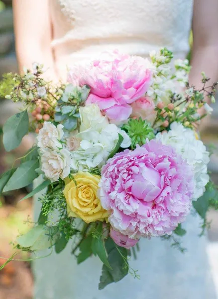 A close-up of a bride holding a bouquet of pink peonies, white roses, and greenery.