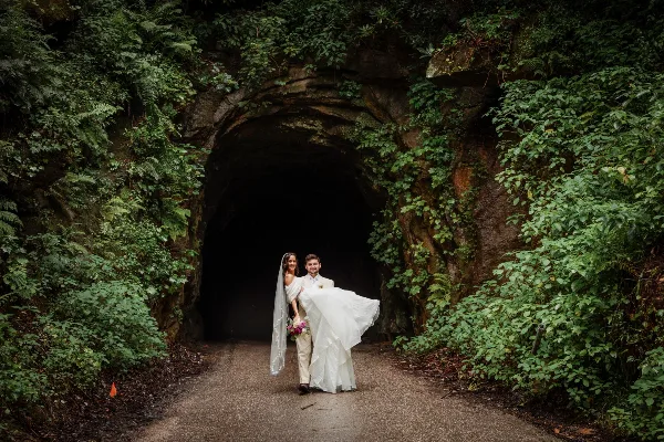 A bride and groom standing together in front of a moss-covered rock archway in a forest setting.
