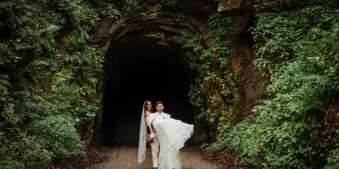A bride and groom standing together in front of a moss-covered rock archway in a forest setting.