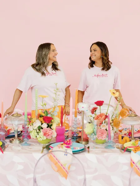 Two women wearing Malyn Made t-shirts standing behind a colorful, decorated wedding table with floral arrangements and candles.
