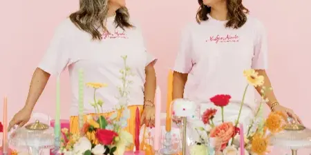 Two women wearing Malyn Made t-shirts standing behind a colorful, decorated wedding table with floral arrangements and candles.