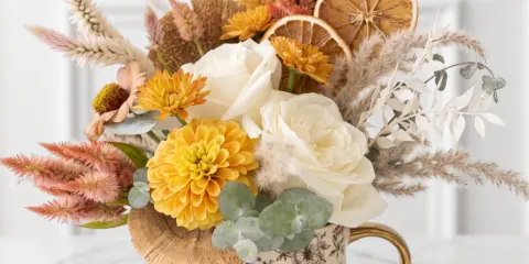 A floral arrangement featuring white roses, yellow dahlias, and dried grasses in a floral-patterned mug with a gold handle.