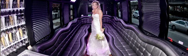 A bride in a white wedding dress standing inside a party bus with purple leather seating and a champagne rack.