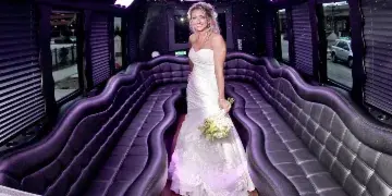 A bride in a white wedding dress standing inside a party bus with purple leather seating and a champagne rack.