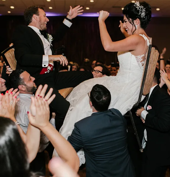 A bride and groom are lifted onto a chair by guests during a lively wedding reception dance party.