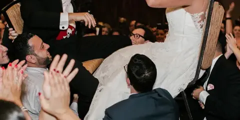 A bride and groom are lifted onto a chair by guests during a lively wedding reception dance party.