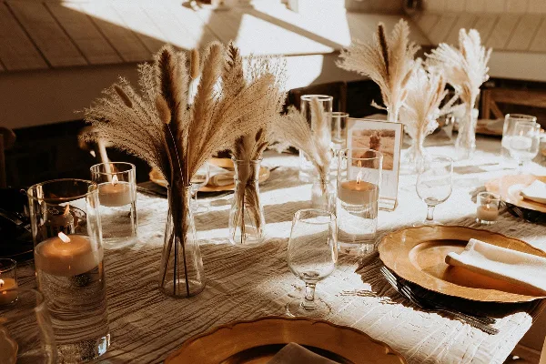 A close-up of an elegant event table setting with pampas grass centerpieces, candles, and gold-rimmed plates.