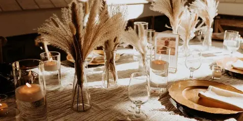 A close-up of an elegant event table setting with pampas grass centerpieces, candles, and gold-rimmed plates.