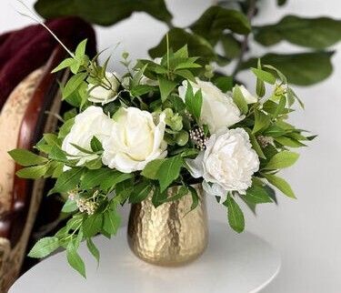 A floral arrangement with white roses and greenery in a gold hammered vase.