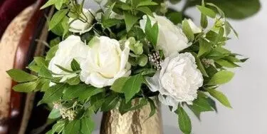 A floral arrangement with white roses and greenery in a gold hammered vase.