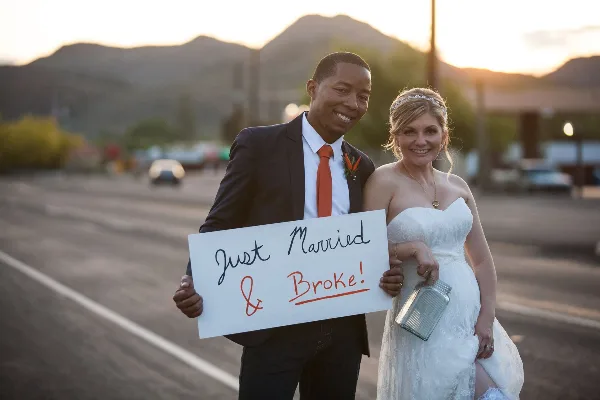 A bride and groom standing outdoors at sunset holding a handwritten 'Just Married & Broke!' sign.