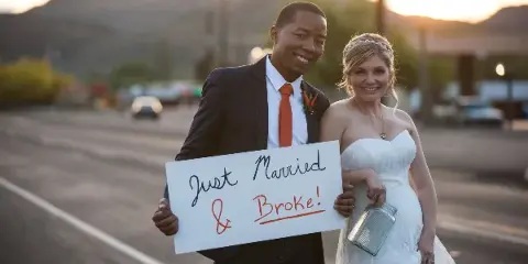 A bride and groom standing outdoors at sunset holding a handwritten 'Just Married & Broke!' sign.